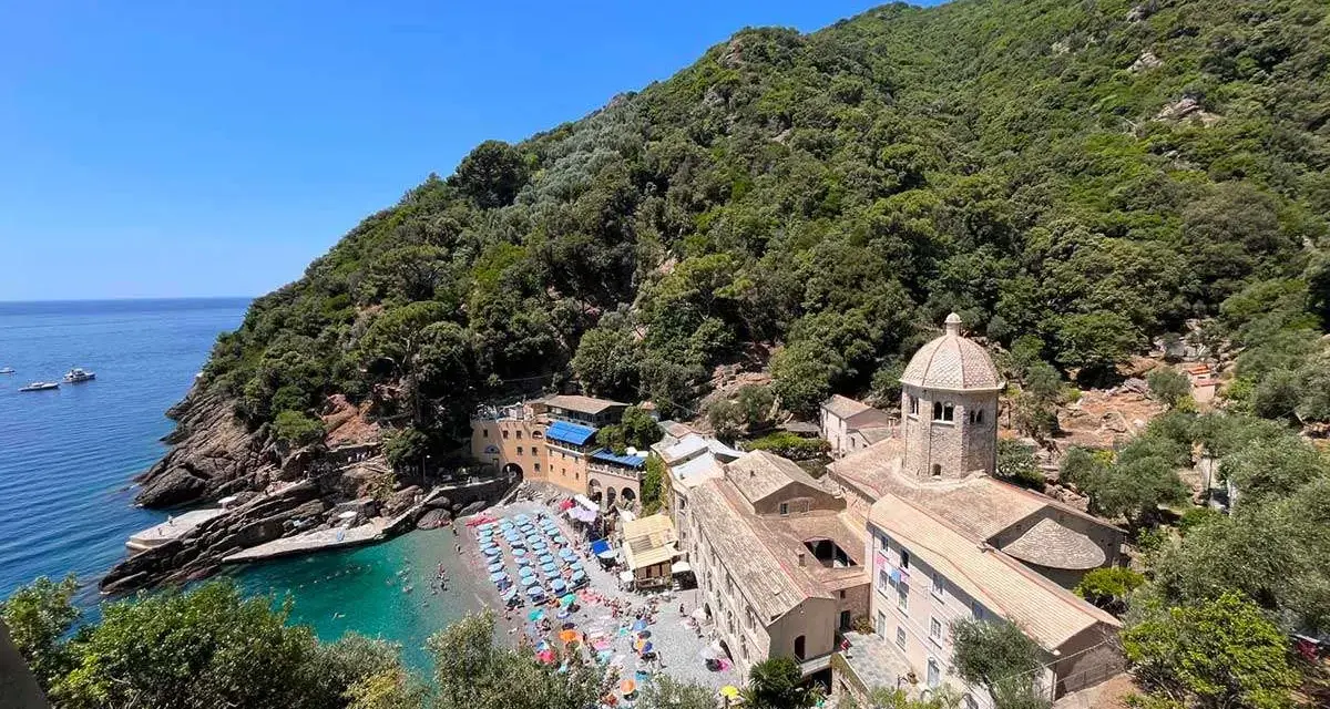 Baia con spiaggia affollata e un antico monastero, un luogo ideale per i cammini in Liguria, con mare azzurro e vegetazione rigogliosa.