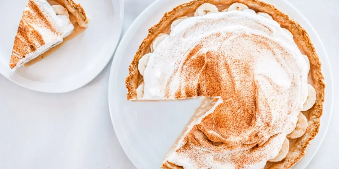 Une part de tarte banoffee traditionnel, garnie de crème fouettée et de cannelle, sur une assiette blanche.
