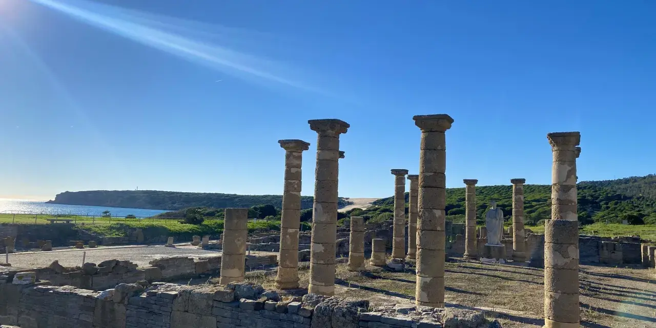 Sunlight streams over ancient roman ruins in Spain, illuminating weathered stone columns and walls against a bright blue sky and distant coastline.