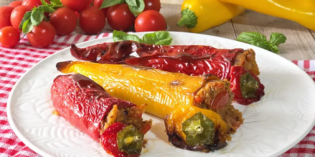 Peperoni cornetti ripieni, rossi e gialli, cotti al forno, su un piatto bianco con pomodorini e basilico.