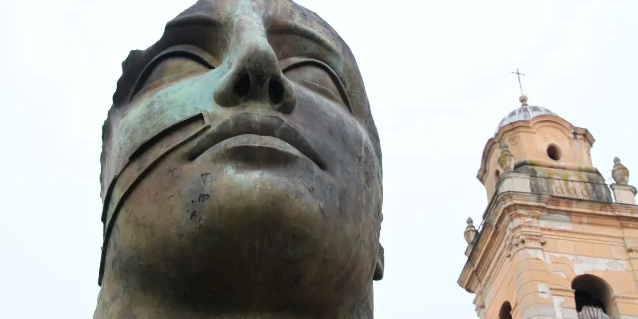 Brązowa, monumentalna rzeźba głowy z widocznym fragmentem dzwonnicy kościelnej w tle.
