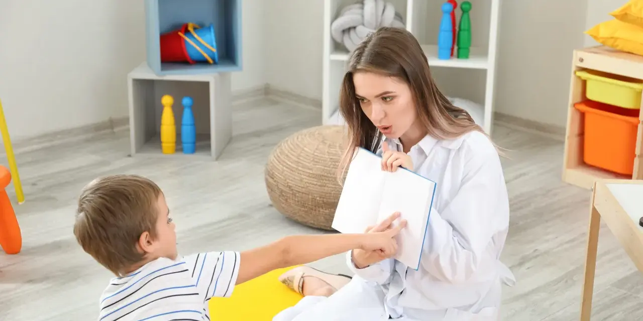 Terapeuta pracuje z dzieckiem, pomagając w rozwoju mowy. Chłopiec wskazuje na książkę, a kobieta uważnie go słucha.