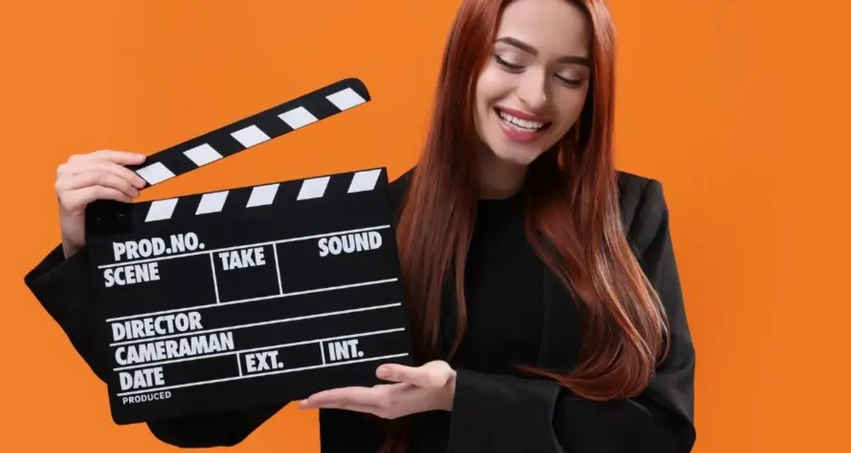 Uśmiechnięta kobieta z rudymi włosami trzyma klaps filmowy. Zastanawiasz się, ile kosztuje hollywood smile w Polsce?