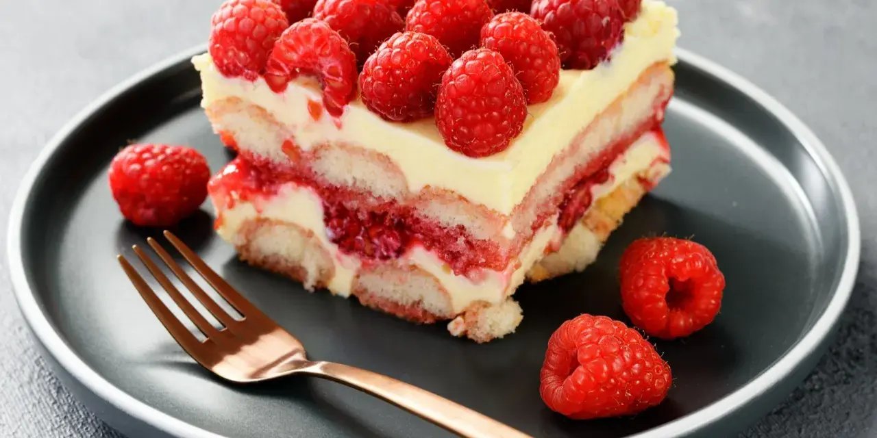 Pyszne tiramisu malinowe z malinami na wierzchu i na talerzu, obok złoty widelec.