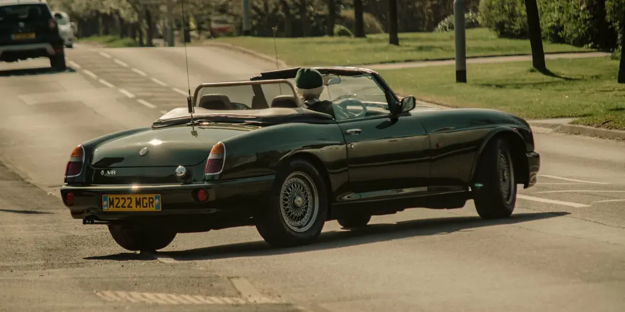 Auto d'epoca verde scuro in movimento, con conducente con cappello. La targa suggerisce che siamo in Inghilterra, dove si guida a destra.