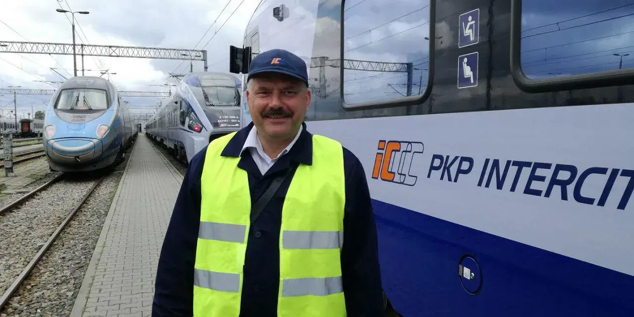 Uśmiechnięty rewident taboru kolejowego w kamizelce odblaskowej stoi obok pociągu PKP Intercity.