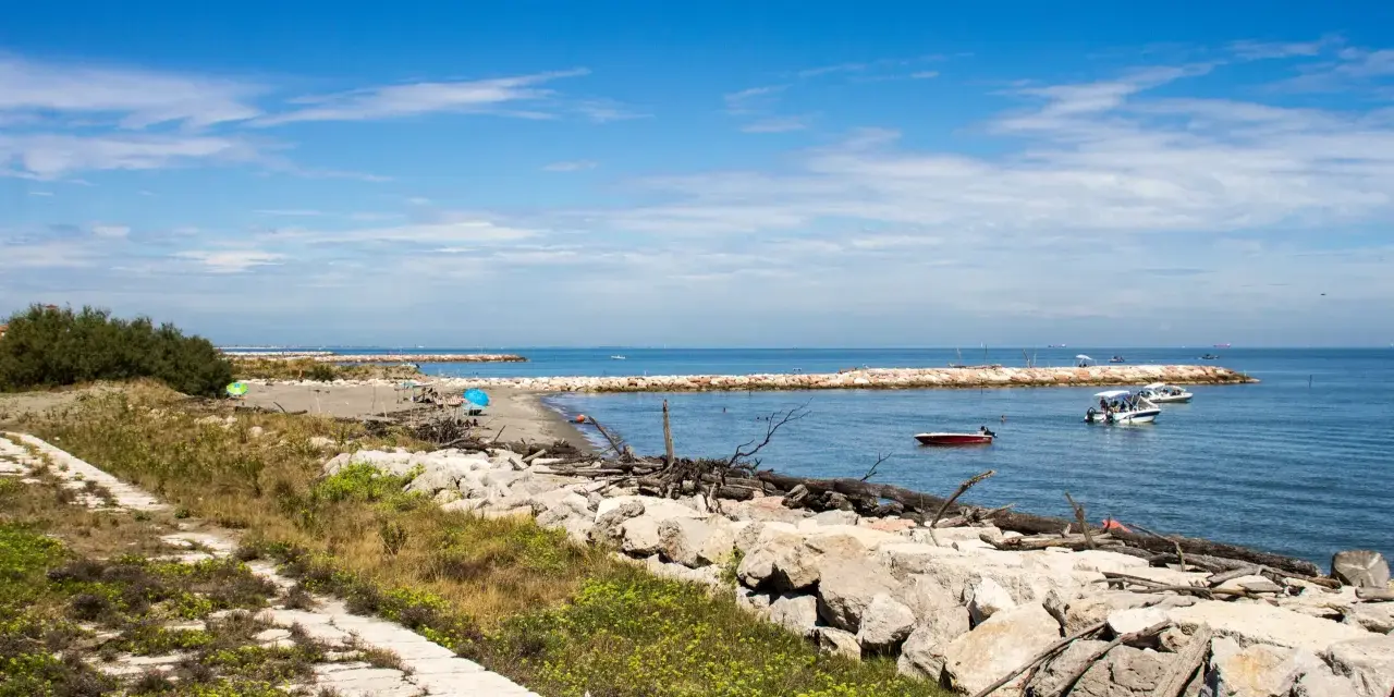 Sentiero in pietra e mare calmo con barche. Per capire come arrivare a Pellestrina da Fusina, immagina di percorrere questa costa.