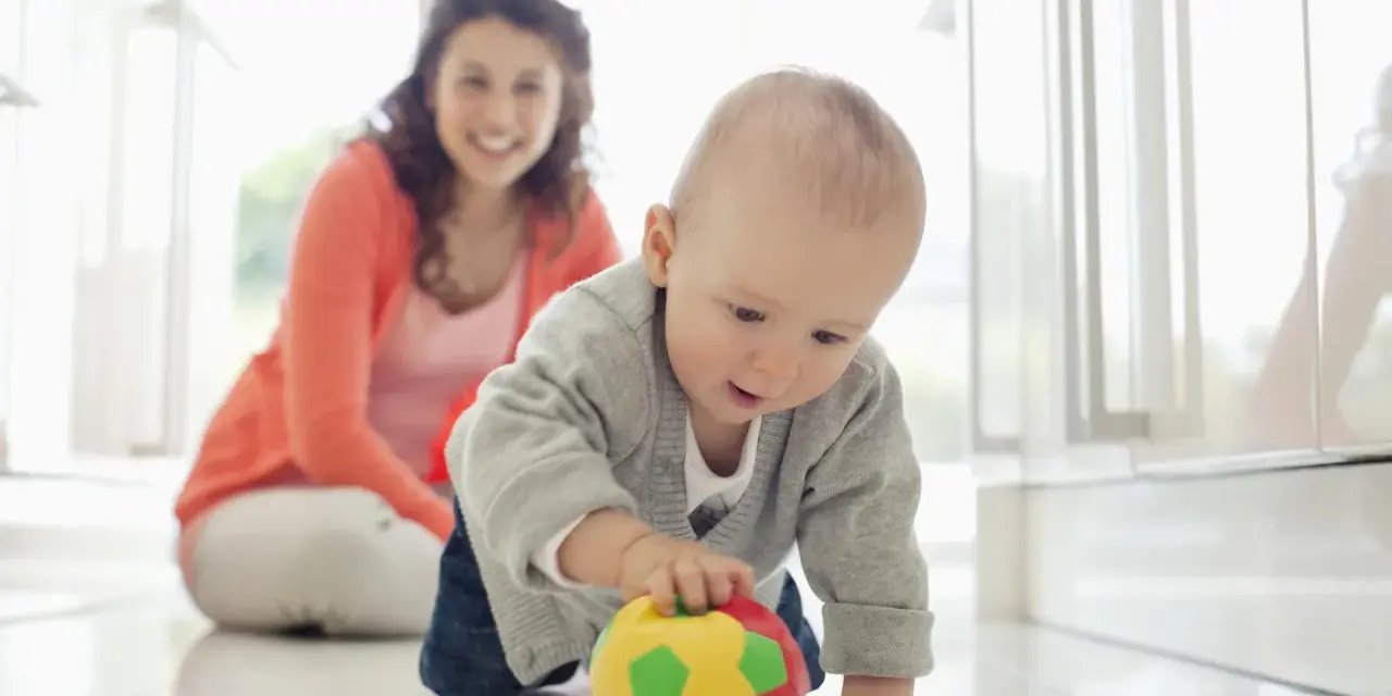 Maluch ćwiczy motorykę dużą, raczkując po podłodze i bawiąc się kolorową piłeczką. Uśmiechnięta mama obserwuje jego postępy.