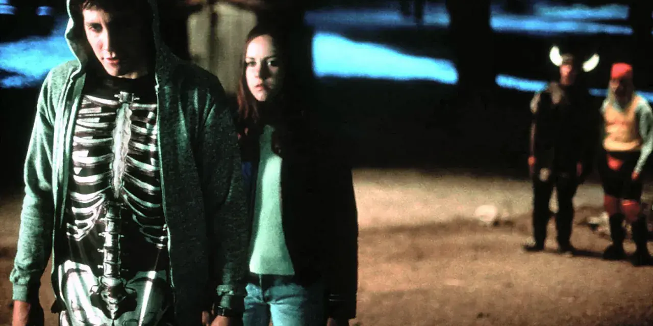 Donnie Darko w bluzie z kapturem i koszulce z nadrukiem szkieletu, obok dziewczyny. W tle dwie postacie w kostiumach.