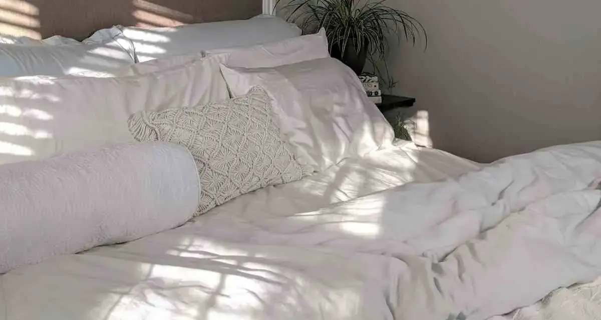 Cama blanca con luz solar filtrada. Aprende como lavar un edredón de plumas para mantenerlo esponjoso.