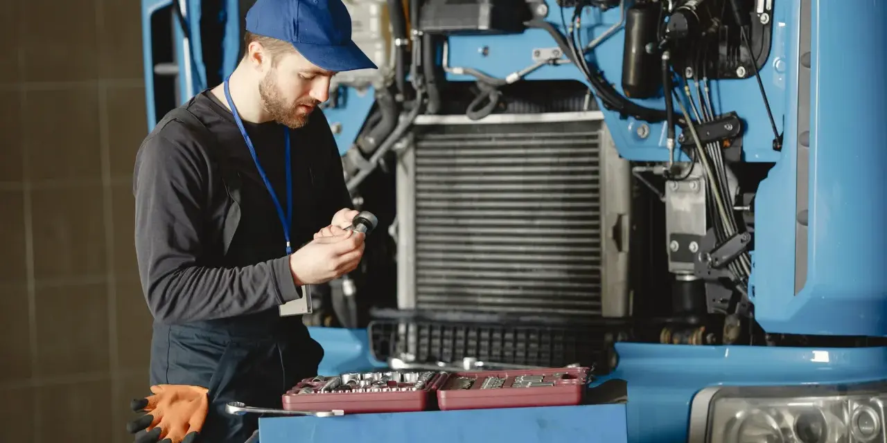 Mechanik w niebieskiej czapce i kombinezonie naprawia ciężarówkę, trzymając narzędzie.