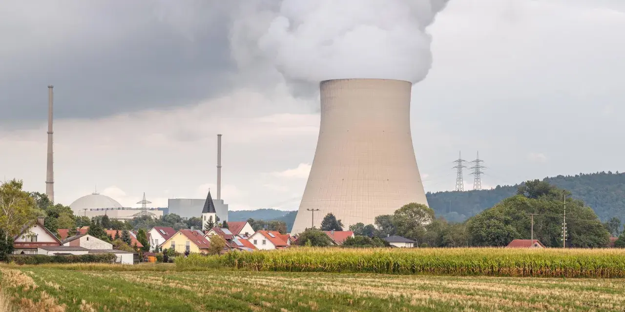 Dampf steigt aus einem Kühlturm eines Atomkraftwerks auf, daneben ein Dorf und ein Maisfeld.