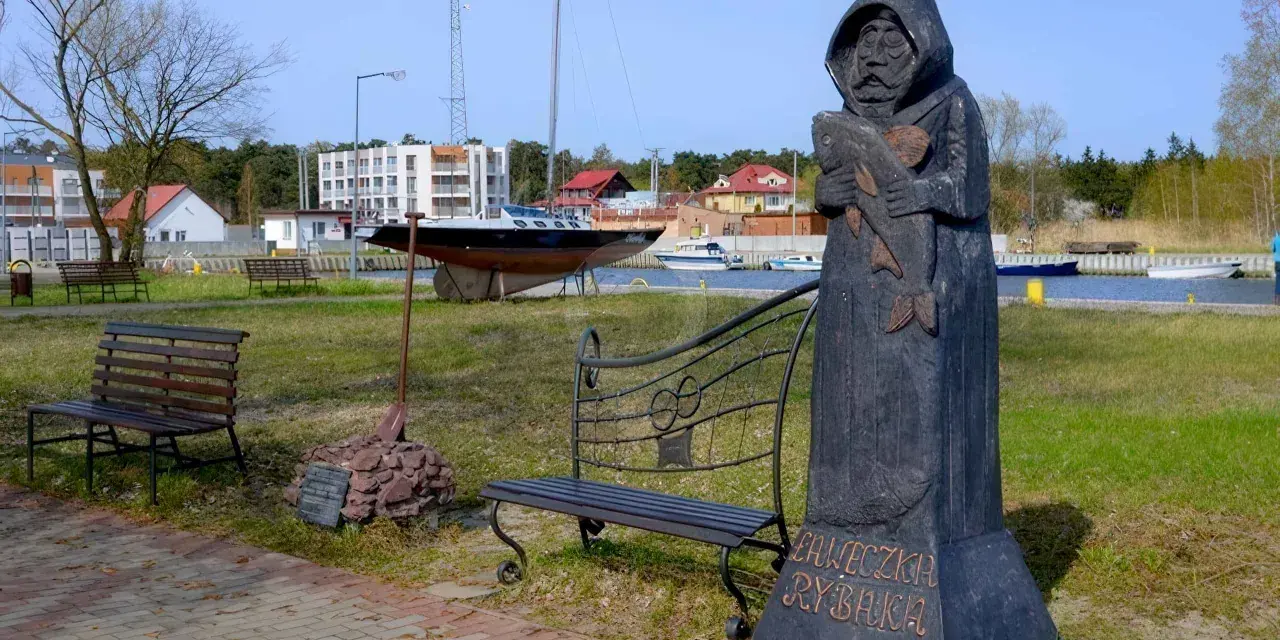 Drewniany pomnik rybaka trzymającego rybę, z napisem "Ławeczka Rybaka". W tle port z łodziami i budynkami.