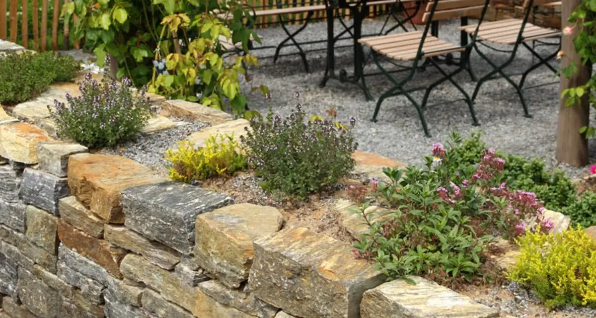 Natürliche Steinmauer mit Kräutern und Blumen, die schöne Ideen für den Garten bietet. Eine Sitzgruppe lädt zum Verweilen ein.