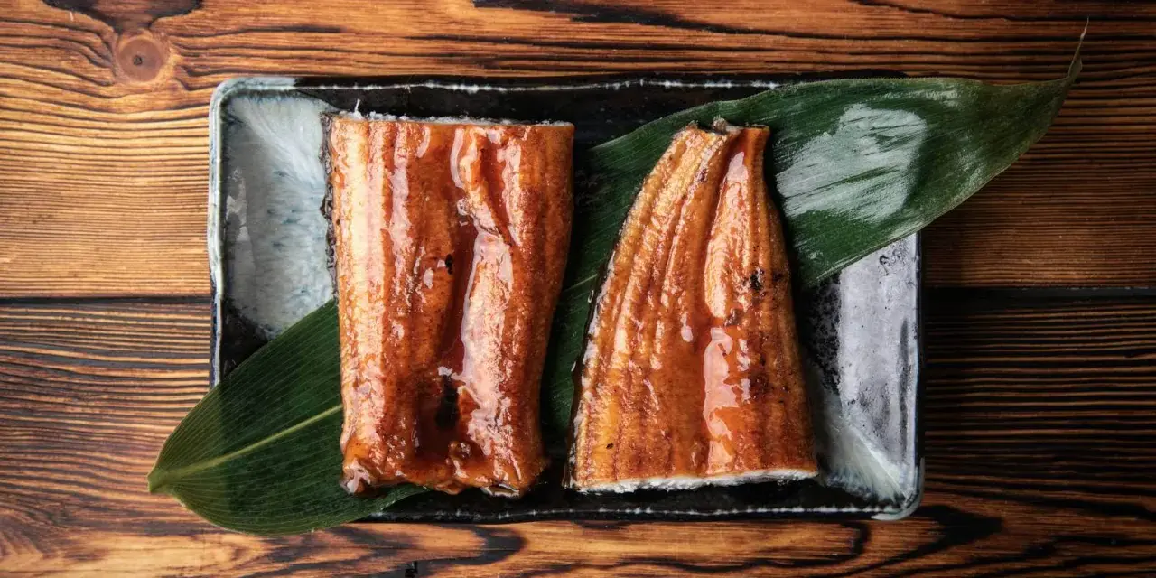 Dwa kawałki glazurowanego węgorza do sushi, podane na liściu na ciemnej zastawie, na drewnianym tle.