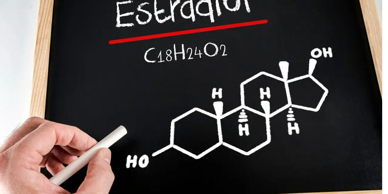 Estradiol co to za badanie? Na tablicy wzór chemiczny estradiolu (C18H24O2) i jego struktura. Ręka z kredą.