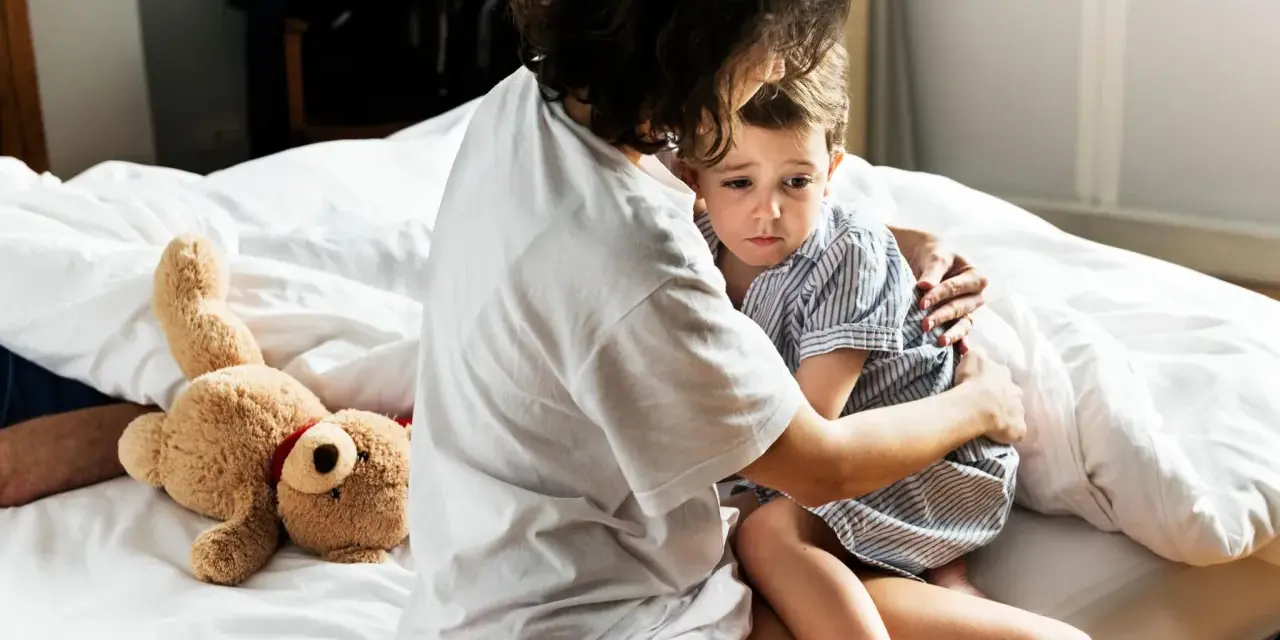Mutter tröstet ihr Kind, das Anzeichen einer Panikattacke bei Kindern zeigt. Ein Teddybär liegt daneben.