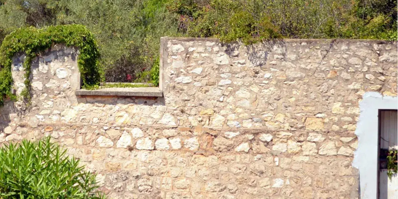 Un mur en pierre extérieur, avec des pierres irrégulières et du mortier, attend de restaurer son charme ancien. La végétation s'y accroche.