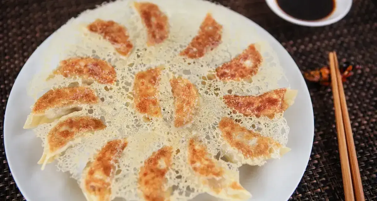 Chrupiące pierożki gyoza ułożone na talerzu, z chrupiącą, złotą panierką. Obok miseczka sosu sojowego i pałeczki.