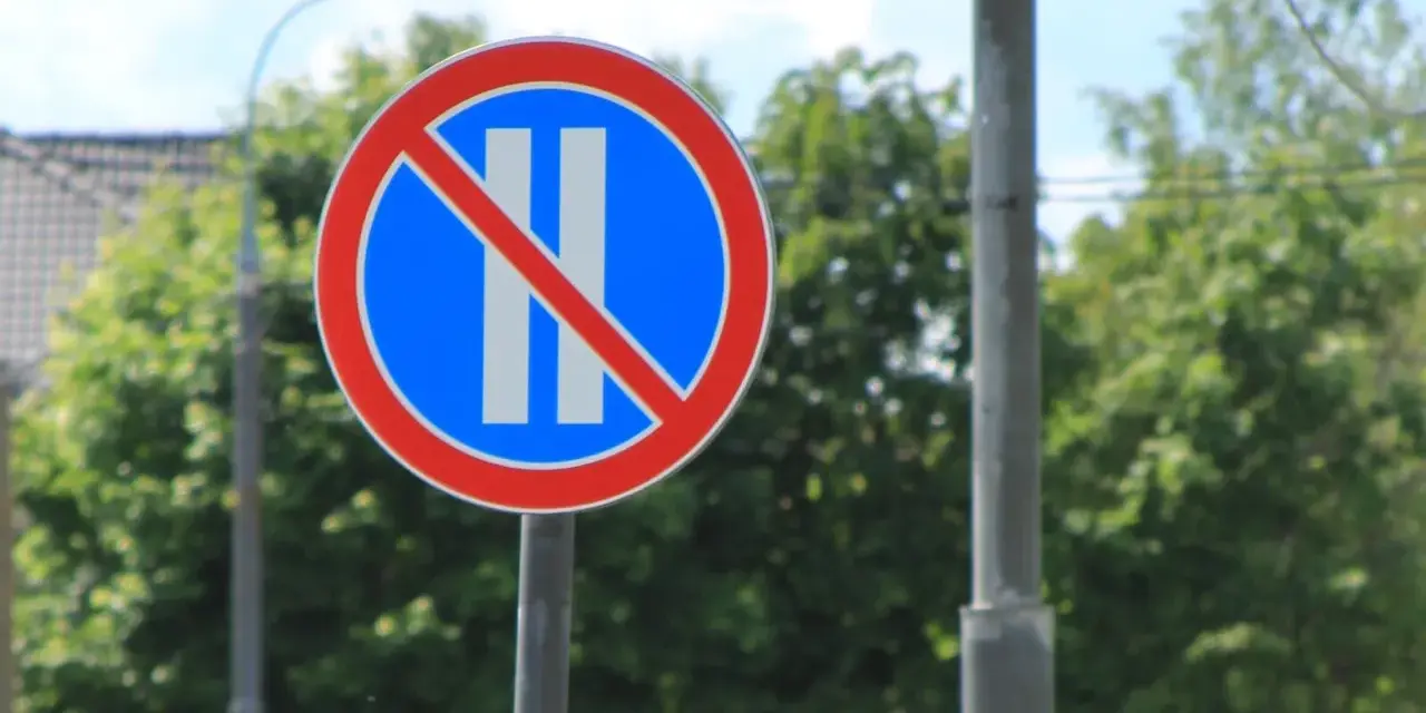 Znak B37 zakazujący postoju. Dwa pionowe białe paski na niebieskim tle, przekreślone czerwoną linią.