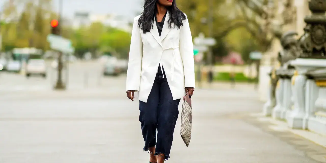 Mujer con estilo **smart casual** camina por la calle con un blazer blanco, jeans oscuros y sandalias de tacón.