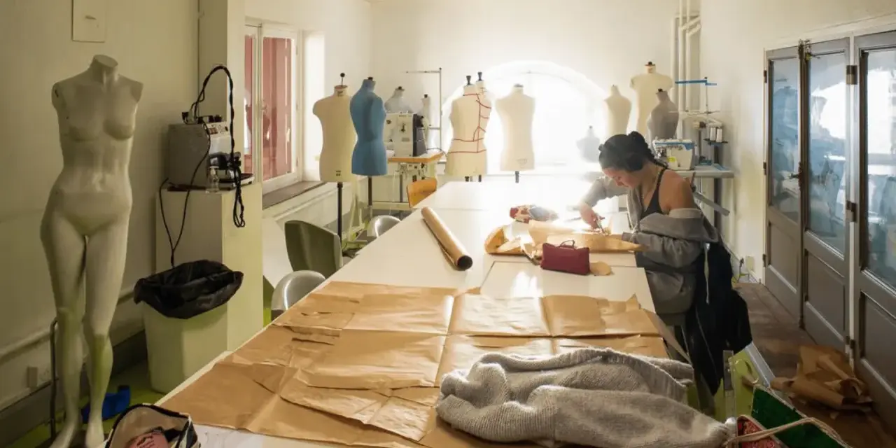 Dans un atelier d'une école d'arts, une jeune femme coupe du tissu sur une grande table. Des mannequins et du matériel de couture sont visibles.