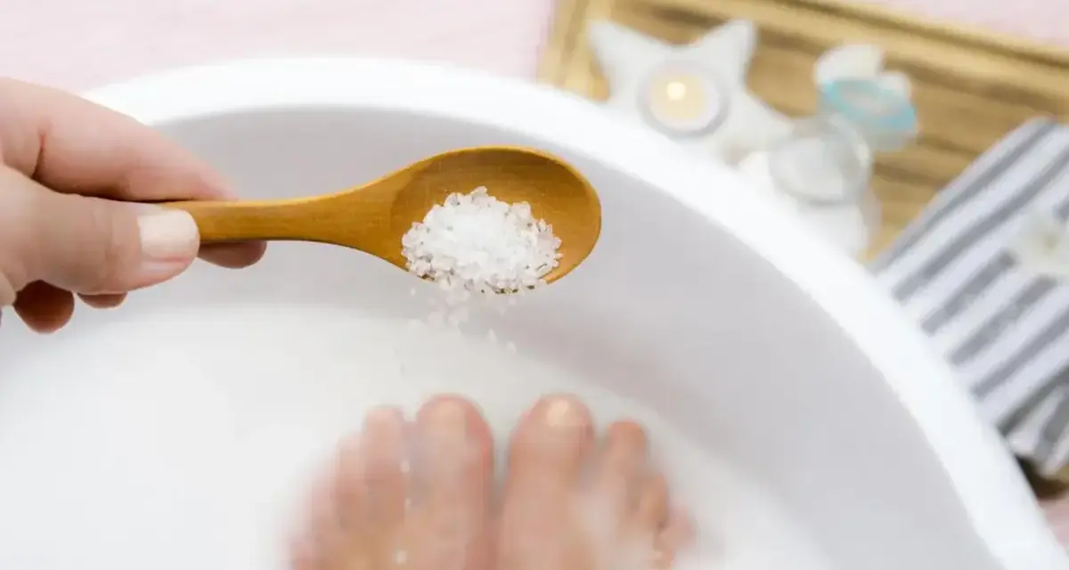 A hand holds a wooden spoon with coarse salt, about to add it to a white basin where feet are soaking. This is a relaxing foot soak with baking soda.