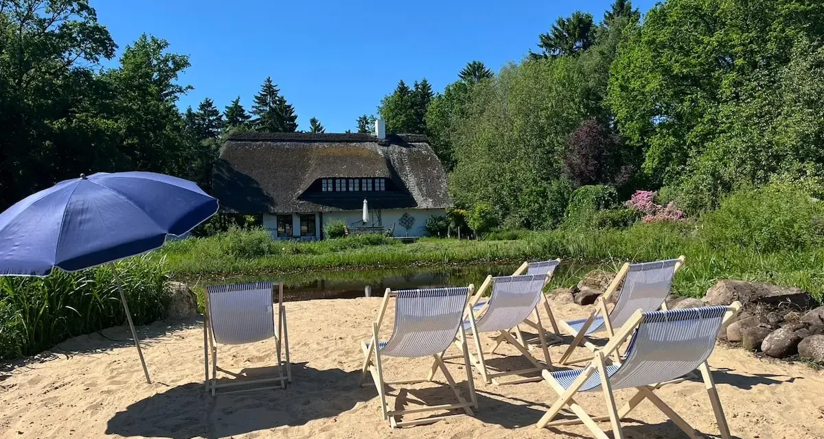 Sonnige Tage am Stedener See: Liegestühle und Sonnenschirm warten auf Besucher vor einem Reetdachhaus.