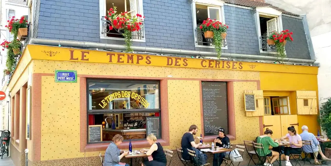 Café "Le Temps des Cerises" con un encantador estilo francés, balcones floridos y gente disfrutando en sus mesas exteriores.