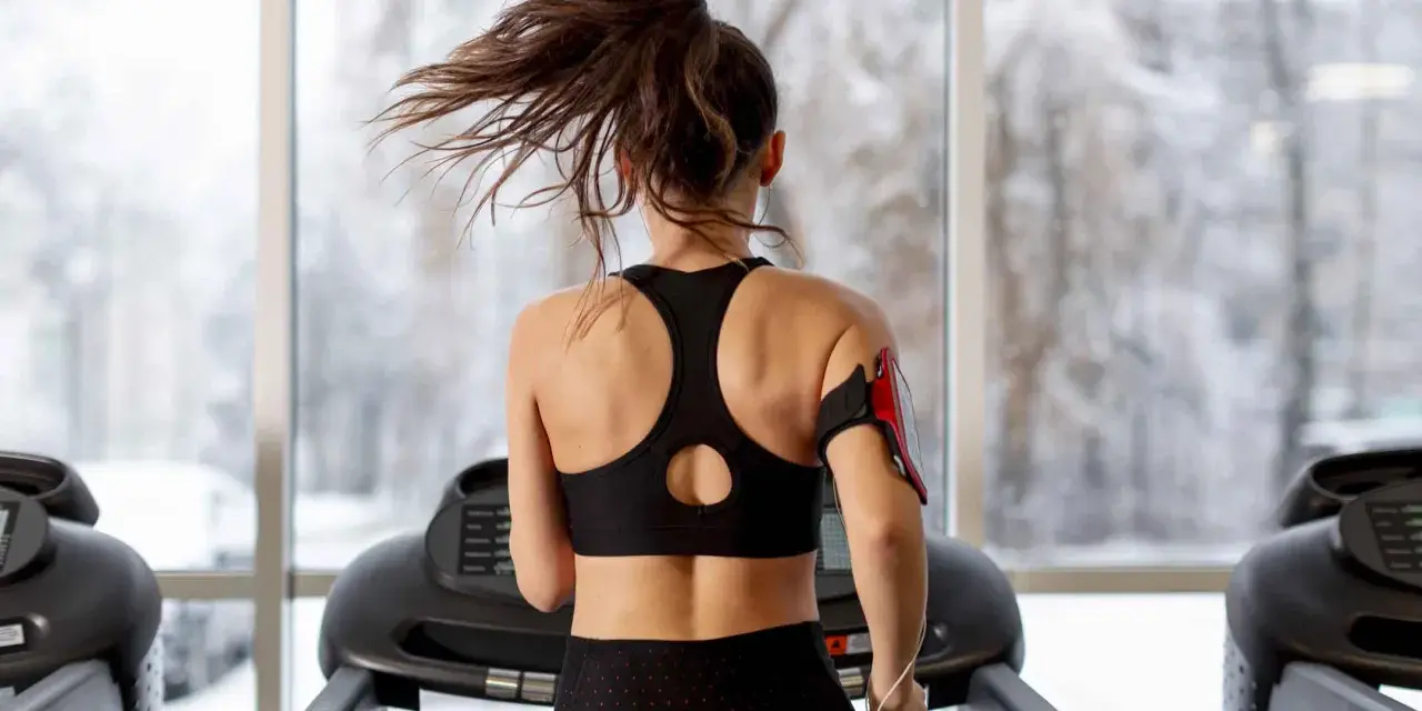 Une femme fait du sport à jeun sur un tapis roulant, ses cheveux flottant au vent. Dehors, la neige tombe.