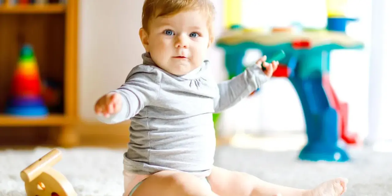 Ein neugieriges 8 Monate altes Baby mit blauen Augen sitzt auf einem flauschigen Teppich und spielt mit Spielzeug.