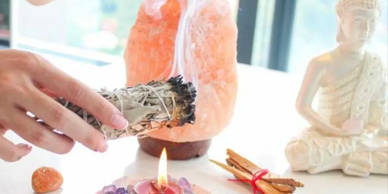 Manos sostienen un manojo de salvia humeante, junto a una lámpara de sal rosa, una vela encendida y una estatua de Buda. Esencial para la limpieza energética de casa.