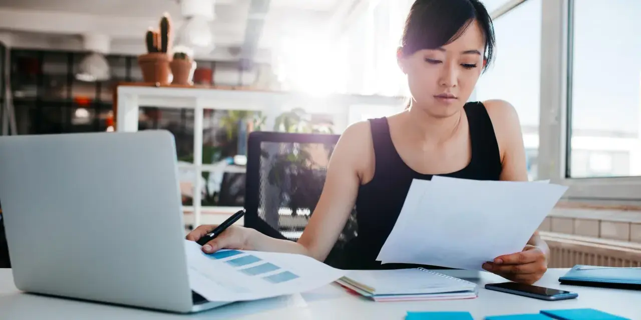 Junge Frau prüft Dokumente, vielleicht die Urlaubsbescheinigung des letzten Arbeitgebers, am Schreibtisch mit Laptop.