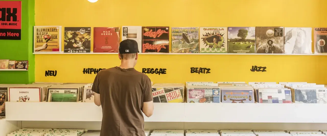 Mann durchstöbert Schallplatten in einem bunten Plattenladen in Berlin. Musikgenres wie HipHop, Reggae, Beatz und Jazz sind angeschrieben.