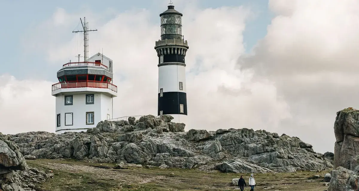 Deux phares se dressent sur une côte rocheuse à Ouessant. Que faire ? Se promener et admirer le paysage, comme ces deux randonneurs.