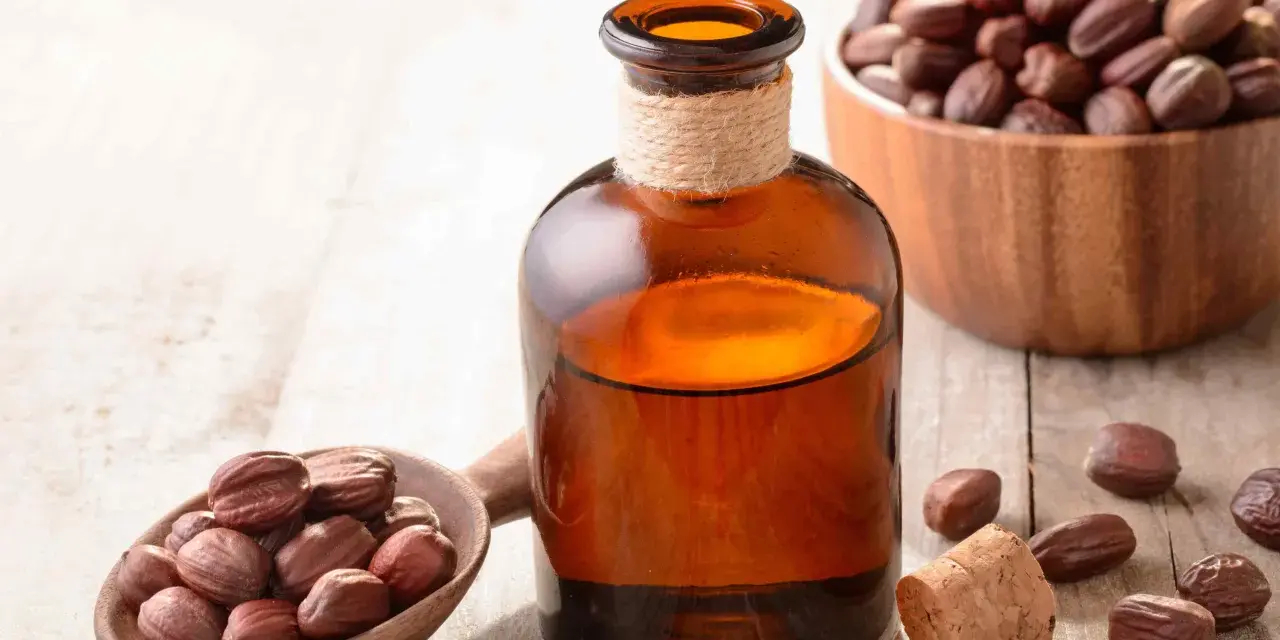 Butelka z bursztynowego szkła z płynnym złotym olejkiem jojoba na włosy, obok drewnianej łyżki z orzechami jojoba.