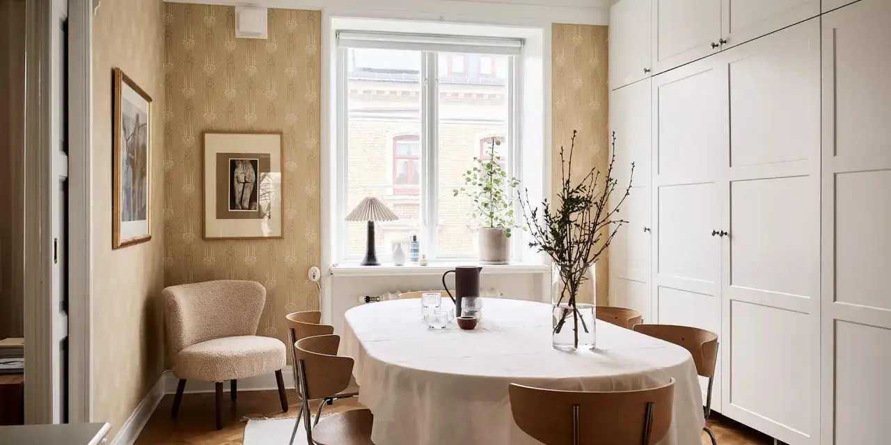Comedor con mesa cubierta, sillas de madera y pared con papel tapiz dorado. Armarios blancos y suelo de parqué.