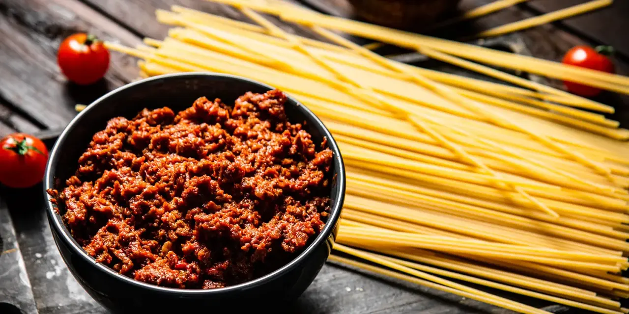 Un tazón de carne para espaguetis a la boloñesa, junto a pasta cruda, tomates y salsa.