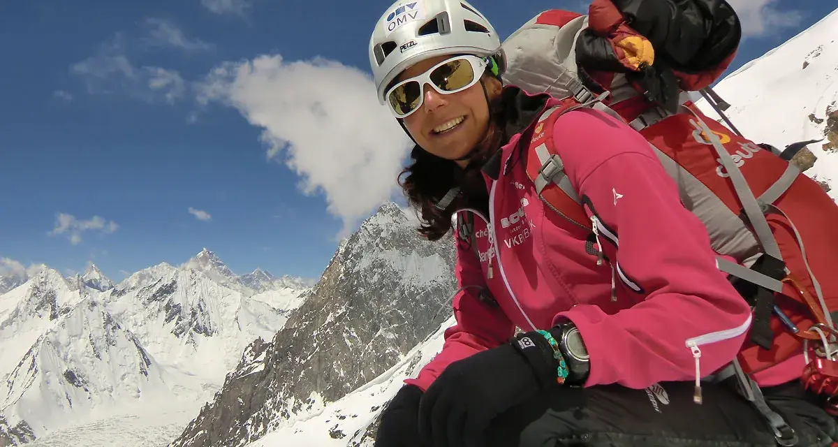 Gerlinde Kaltenbrunner, lächelnd in voller Bergsteigerausrüstung, vor einer majestätischen Schneelandschaft.