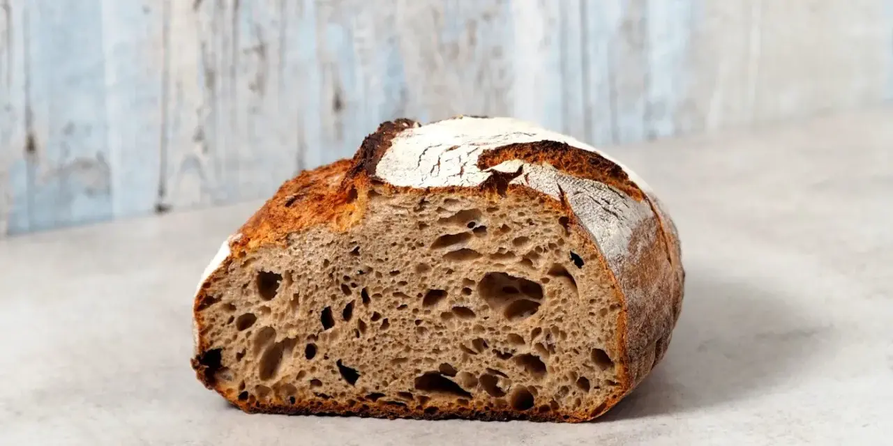 Ein halbes, knuspriges schnelles Sauerteigbrot mit großer Krume und rustikaler Kruste liegt auf einem hellen Untergrund vor einer hellblauen Holzvertäfelung.