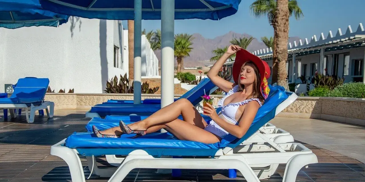 Kobieta w białym bikini i czerwonym kapeluszu relaksuje się na leżaku z drinkiem, ciesząc się wakacjami w stylu ultra all inclusive.