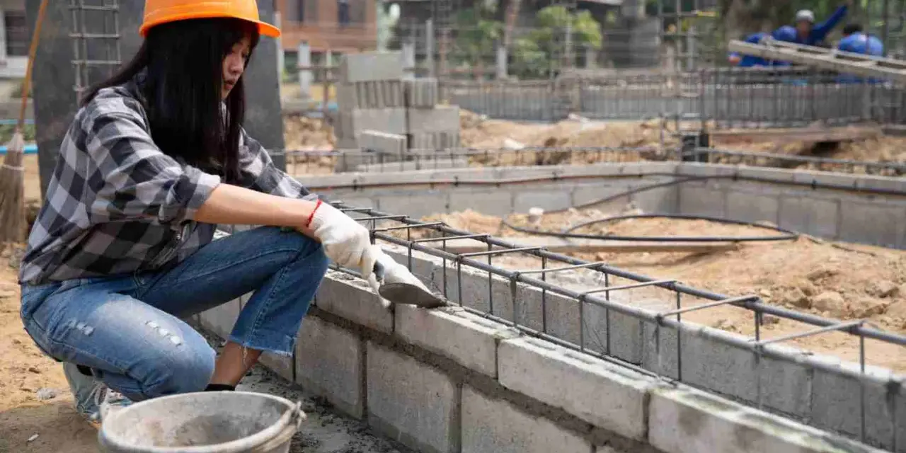 Une ouvrière en casque orange pose du ciment sur un mur en parpaing. Le chantier progresse.