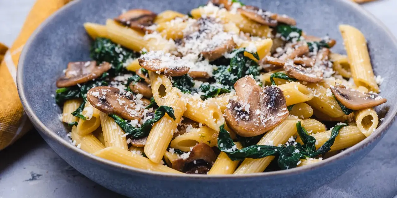 Penne z pieczarkami i szpinakiem, posypane parmezanem, w szarej misce.