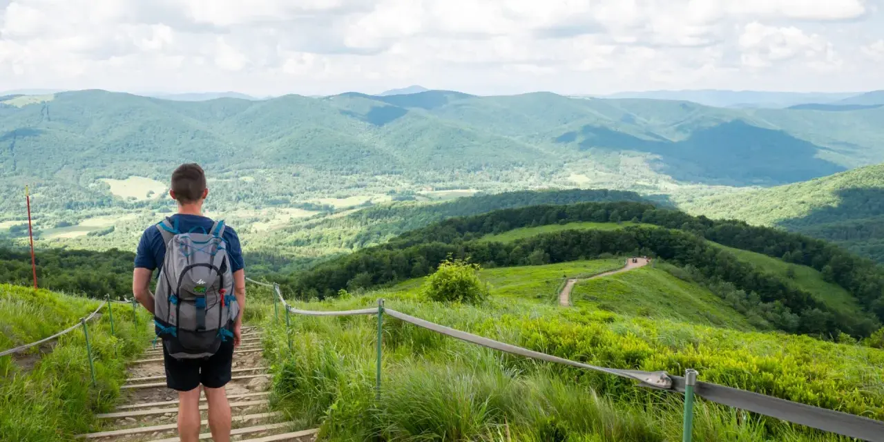 Mężczyzna z plecakiem schodzi po kamiennych schodach, podziwiając rozległe, zielone **Bieszczady góry**.
