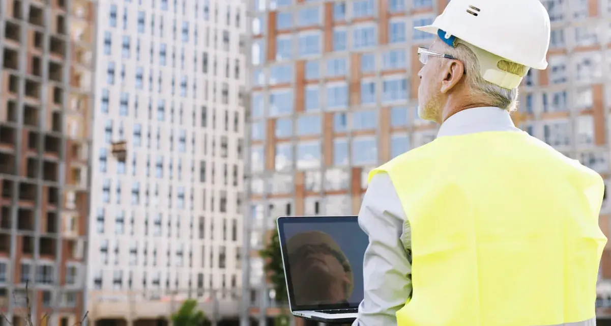 Ingeniero con casco y chaleco reflectante revisa un portátil frente a edificios en construcción. A pesar de un posible **ite desfavorable**, su mirada es decidida.