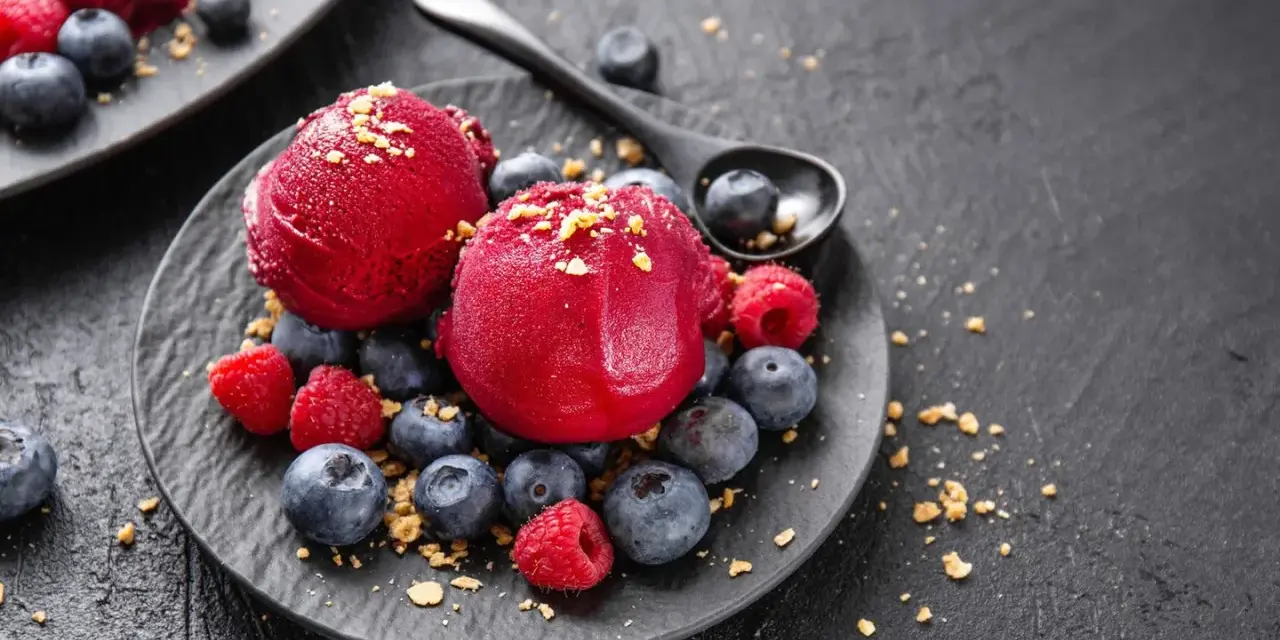 Zwei Kugeln Campari Sorbet mit frischen Beeren und Streuseln auf einem schwarzen Teller.