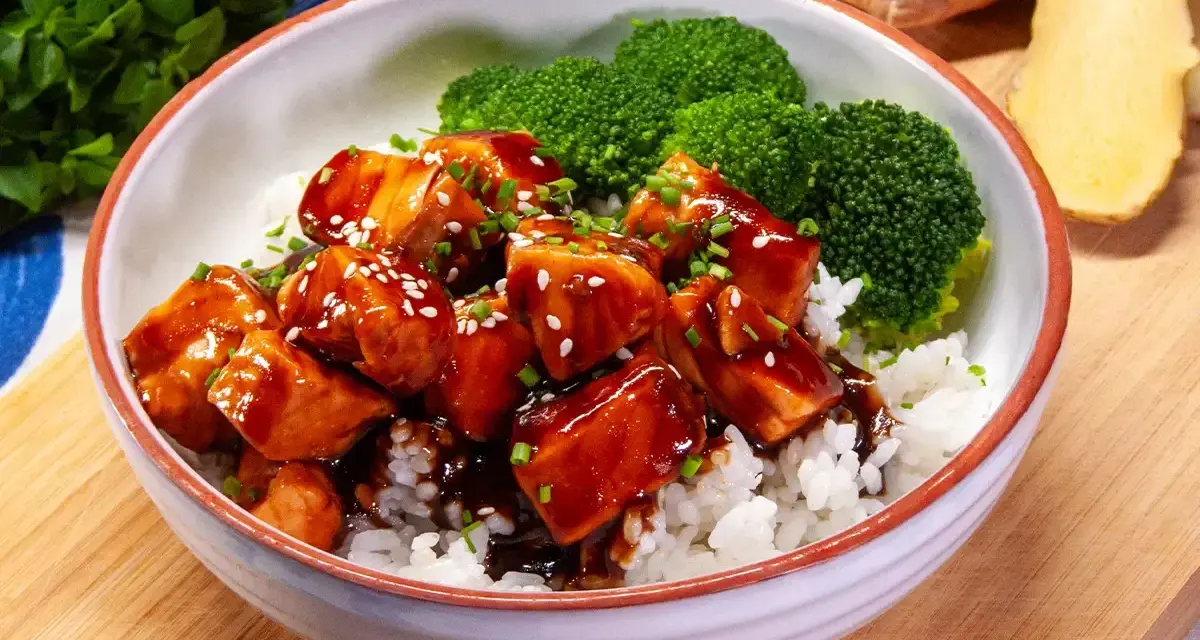 Miseczka ryżu z kawałkami lśniącego lososia teriyaki, posypanego sezamem i szczypiorkiem, obok parujących różyczek brokułów.