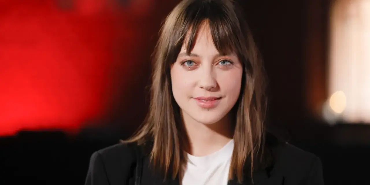 Maria Sobocińska z niebieskimi oczami i grzywką, ubrana w czarną marynarkę i białą koszulkę, uśmiecha się lekko na tle czerwonego światła.