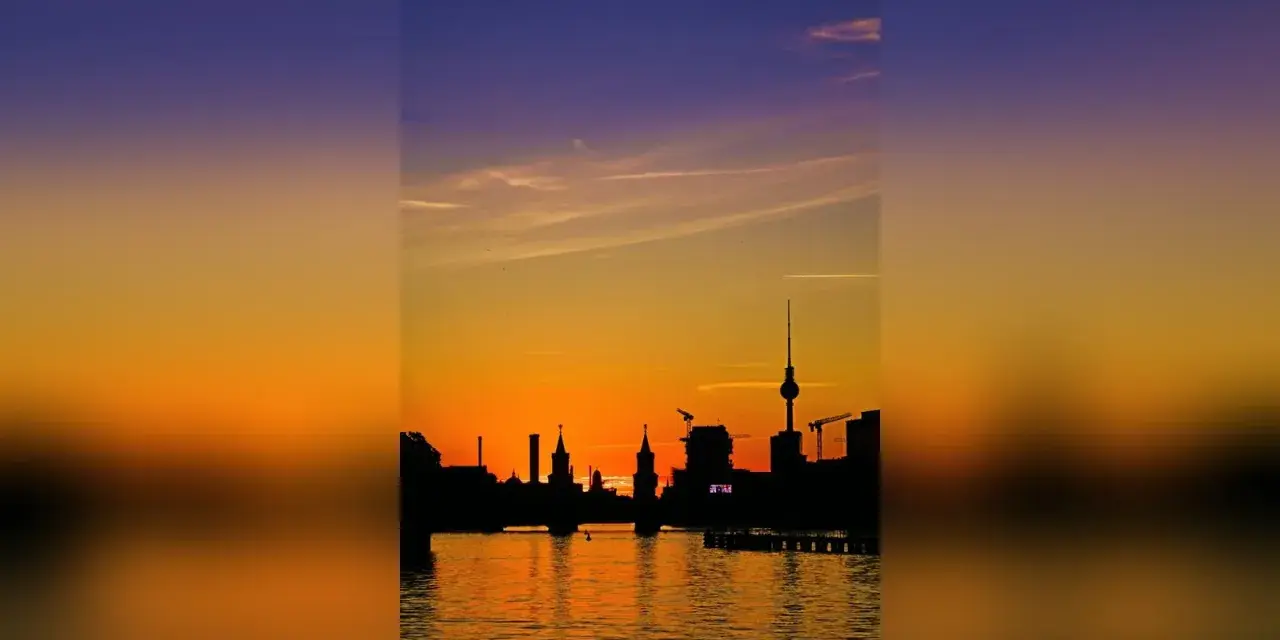 Silhouetten der Berliner Skyline spiegeln sich im Wasser. Ein dramatischer Sonnenuntergang taucht den Himmel in Orange- und Violetttöne.