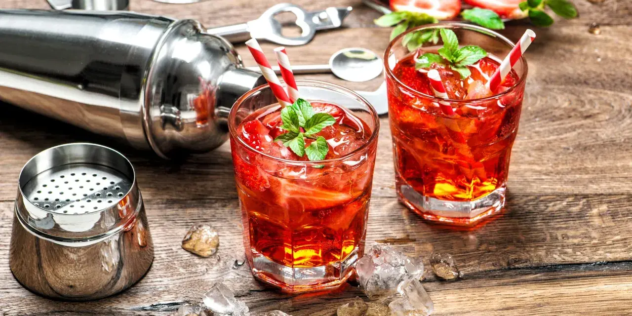 Zwei erfrischende Erdbeer-Cocktails mit Minze und Eiswürfeln, bereit zum Genießen. Ein Shaker und Bar-Werkzeuge deuten auf das kunstvolle Alkohol mischen hin.
